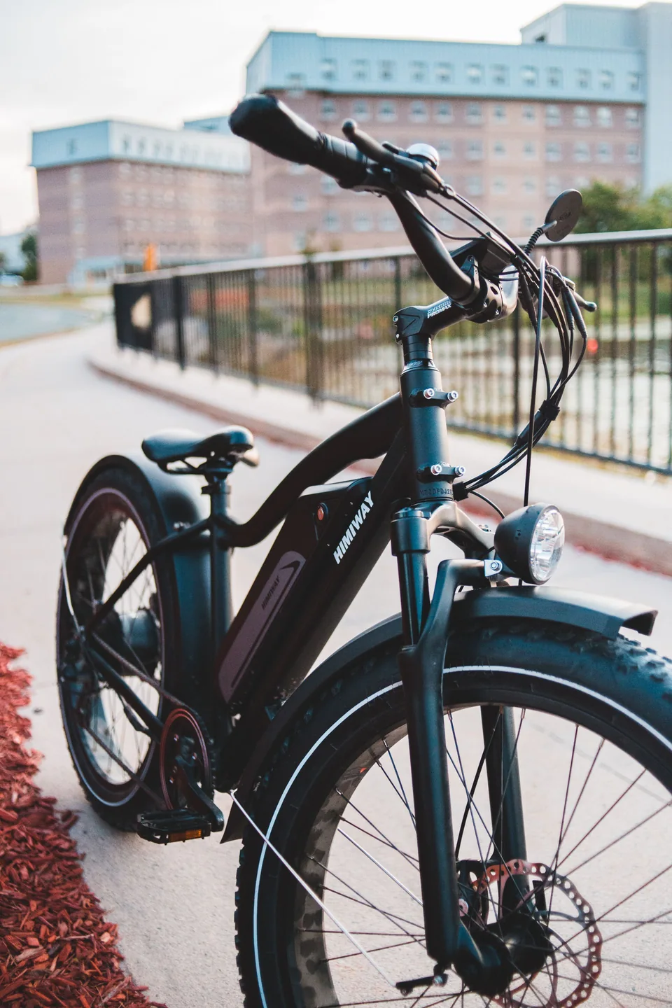 Black electric bicycle beside an urban waterfront; lifestyle imagery, not a catalogue product